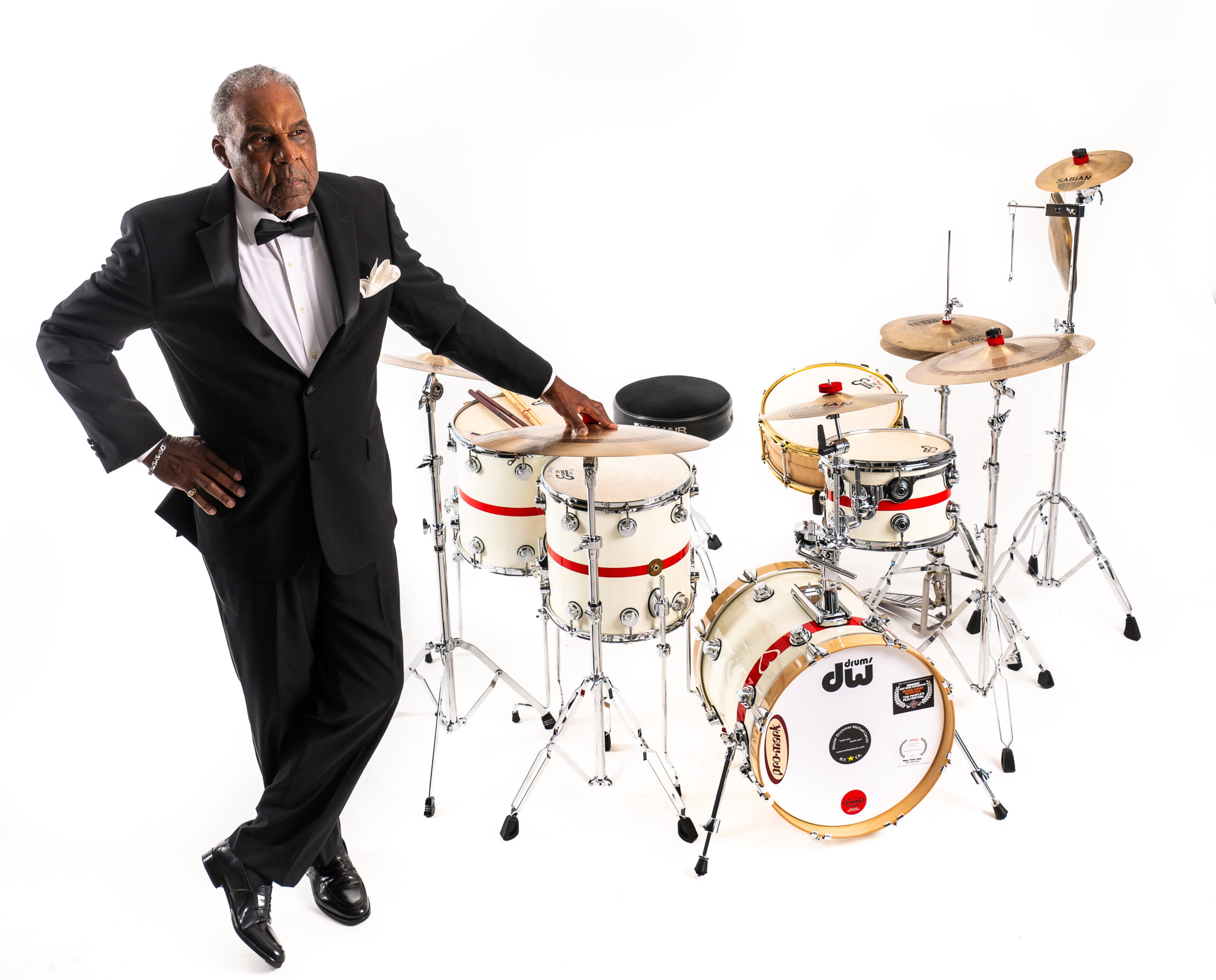 Michael Carvin in a black suit and bow tie is confidently posing next to a white and red drum set against a white background.